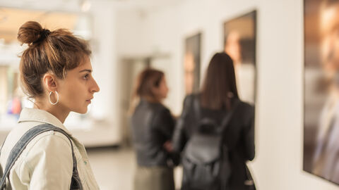 Am linken Bildrand ist eine Frau mit einem Dutt, in einer hellen Jacke und mit einer Tasche über der Schulter zu sehen, die sich ein Gemälde ansieht, dass am rechten Bildrand angeschnitten ist. Im Hintergrund sind zwei weitere Frauen in dunklen Jacken zu sehen, die sich andere Gemälde ansehen.