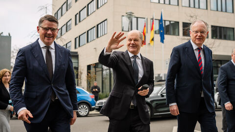 Drei Männer im Anzug laufen über eine Straße. Im Hintergrund ist ein Haus zu sehen. Der Mann in der Mitte winkt.