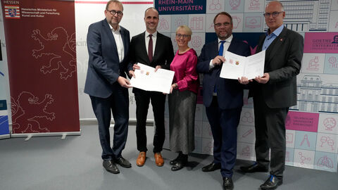 Fünf Personen stehen nebeneinander und halten zwei Urkunden hoch.